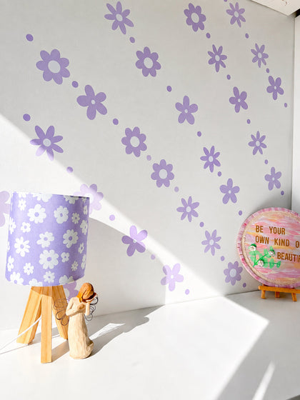 A styled corner with a white wall decorated with lilac decal sticker flower. A lavender lamph white flowers sits on a small wooden tripod table, next to a wooden figurine and a round sign that reads 'BE YOUR OWN KIND OF BEAUTY'.