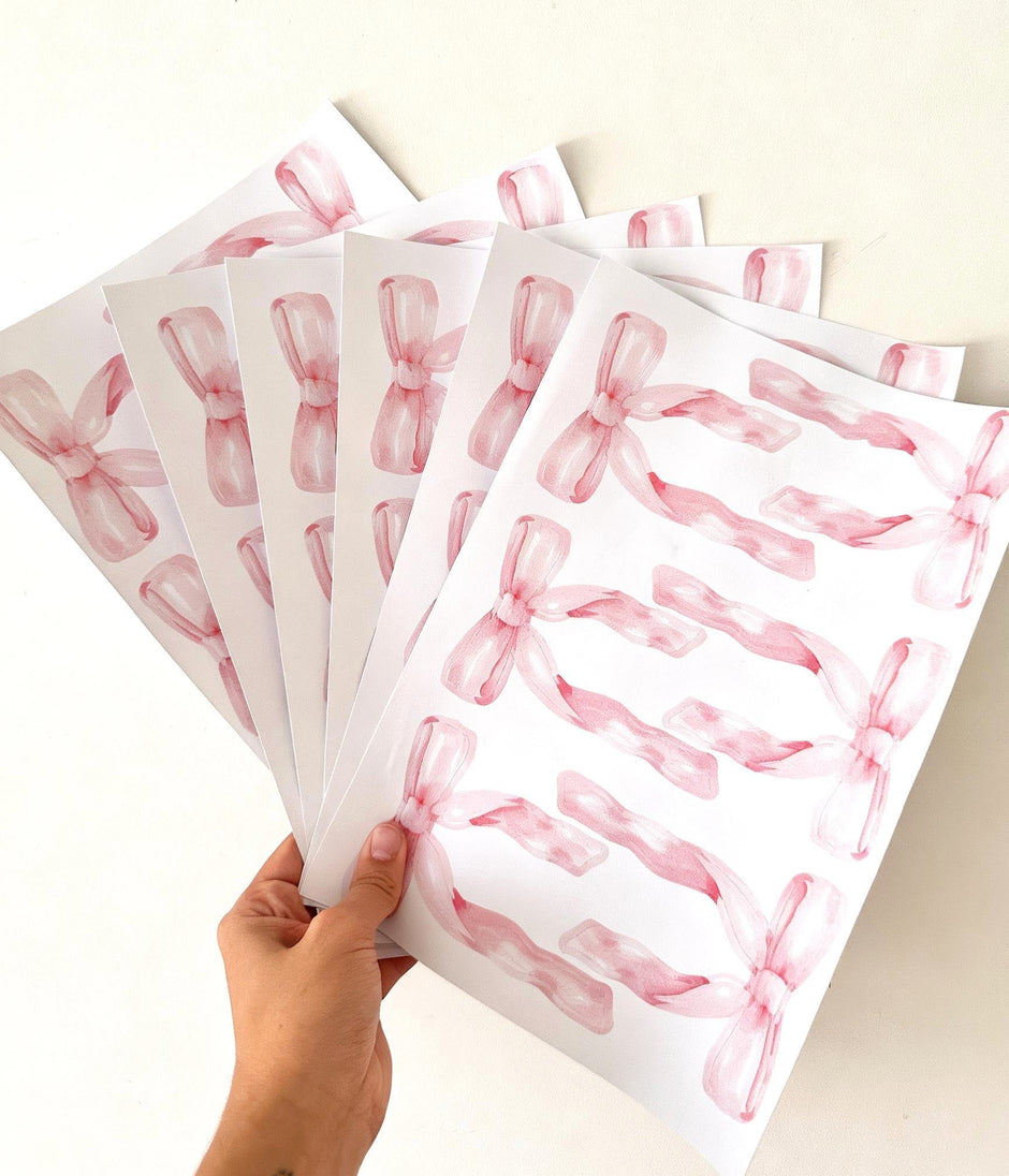 A hand holds a fan of white sheets printed with pink watercolor ribbon reusable wall decal stickers on a plain light background.