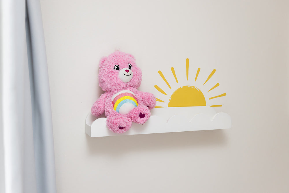 Pink teddy bear with a rainbow on a white shelf with a reusable yellow sun wall decal sticker against a light gray wall.