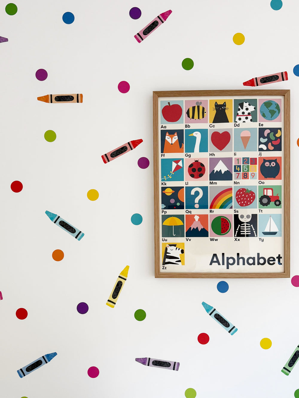 A framed alphabet children's poster with colorful illustrated letters and words, displayed on a white wall surrounded by scattered crayon-and bright dot reusable dots brightly coloured wall paper stickers
