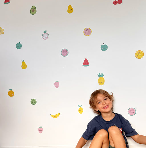 A smiling young boy sits on a white floor against a white wall decorated with colorful, reusable cute fruit wall decal stickers.