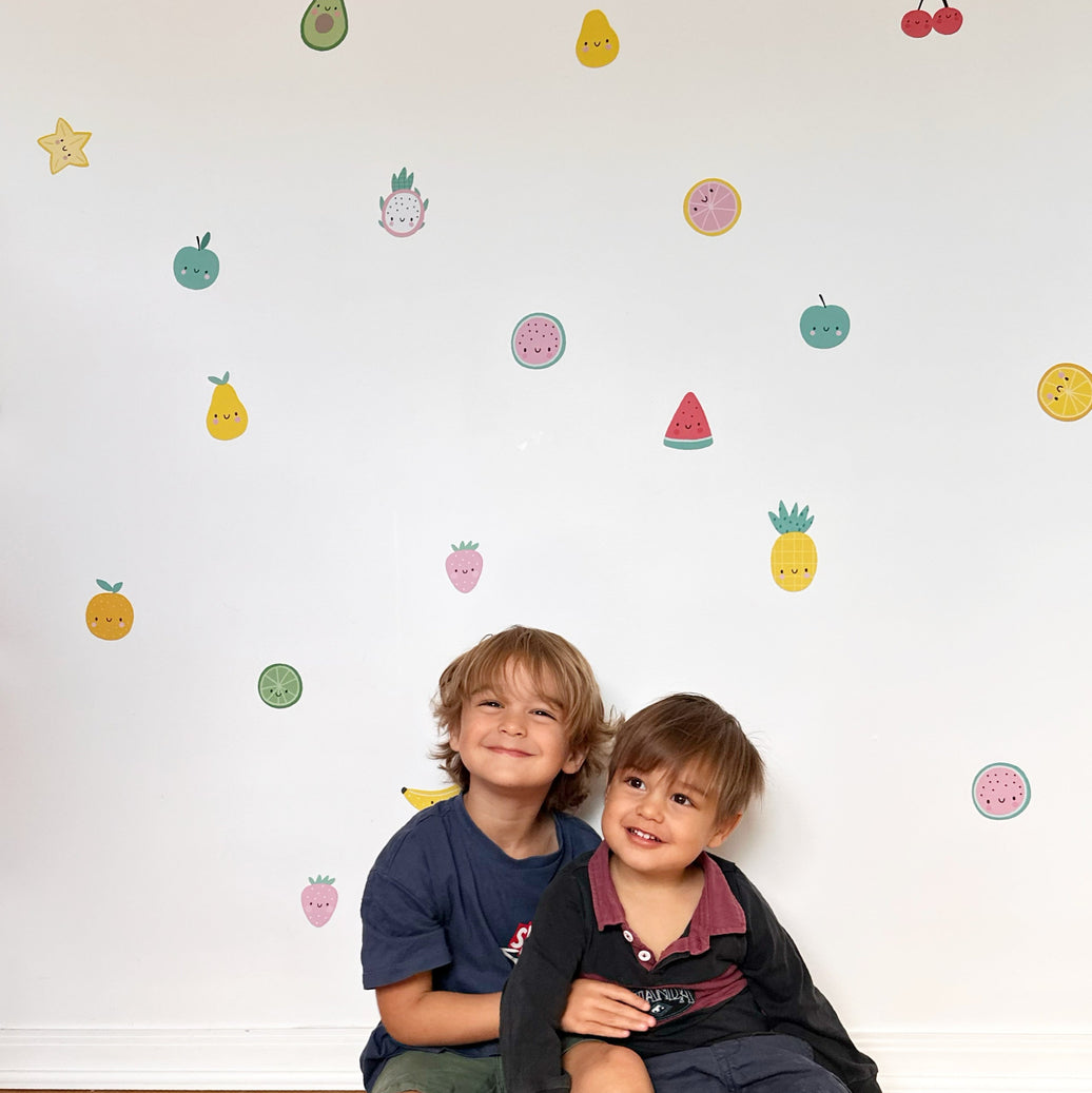 Two young boys sitting on a light wood floor in front of a white wall decorated with colorful reusable fruit and avocado wall decal stickers.