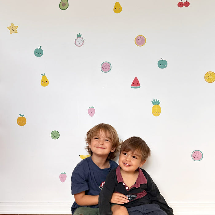Two young boys sitting on a light wood floor in front of a white wall decorated with colorful reusable fruit and avocado wall decal stickers.