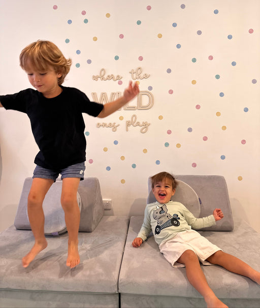 Two children playing on a gray foam play couch in a child-friendly room with pastel polka dot wall decal stickers and the phrase 'where the wild ones play' on the wall.