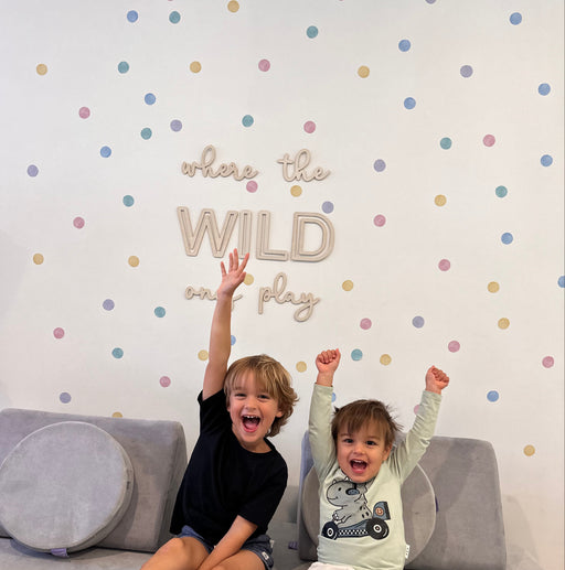 Two young children sitting on a gray sofa in front of a white wall decorated with pastel polka dot wall decal stickers and the words 'where the WILD ones play'. 