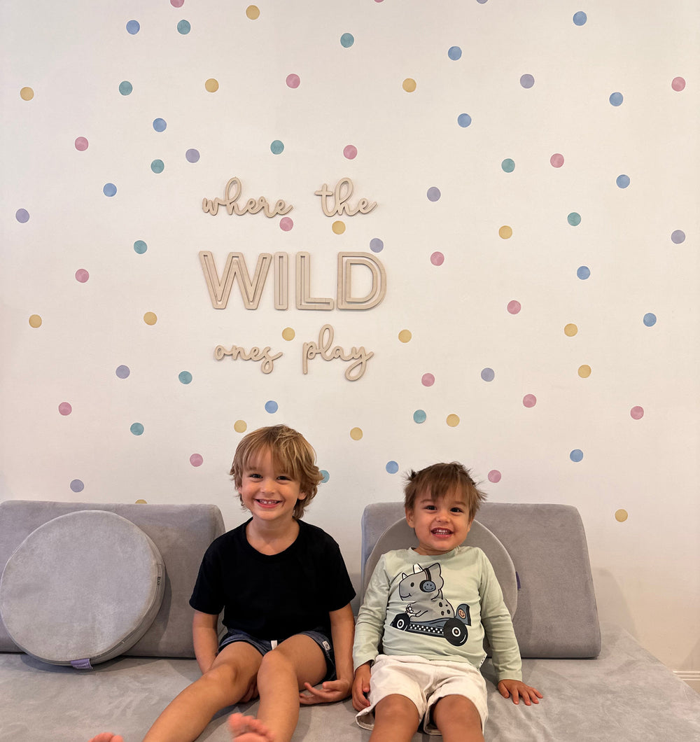 Two young boys sitting on a gray sofa in front of a white wall decorated with pastel polka dot reusable walldecal stickers and the phrase 'where the WILD ones play'.