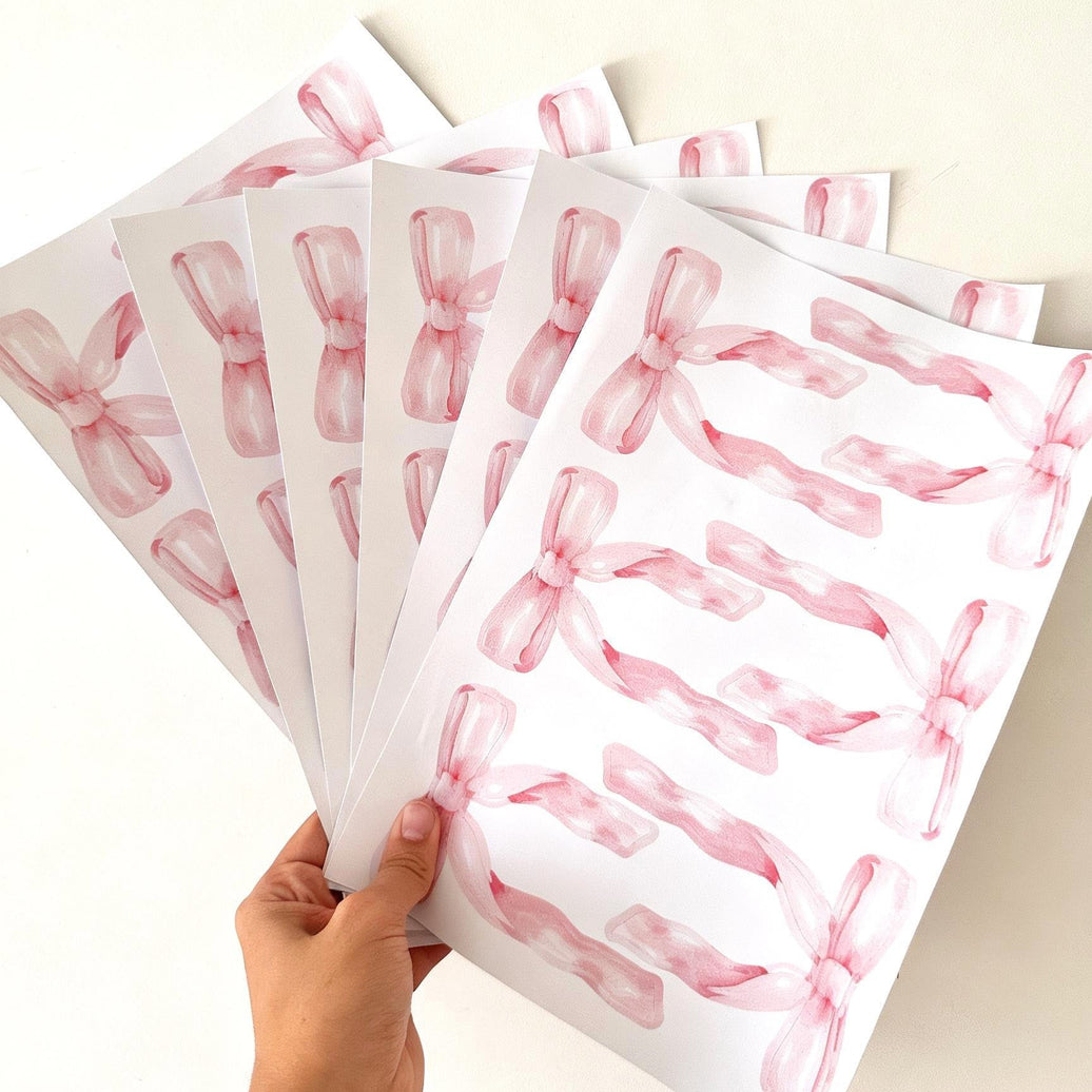A hand holds a fan of white sheets printed with pink watercolor ribbon reusable wall decal stickers on a plain light background.