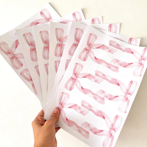 A hand holds a fan of white sheets printed with pink watercolor ribbon reusable wall decal stickers on a plain light background.